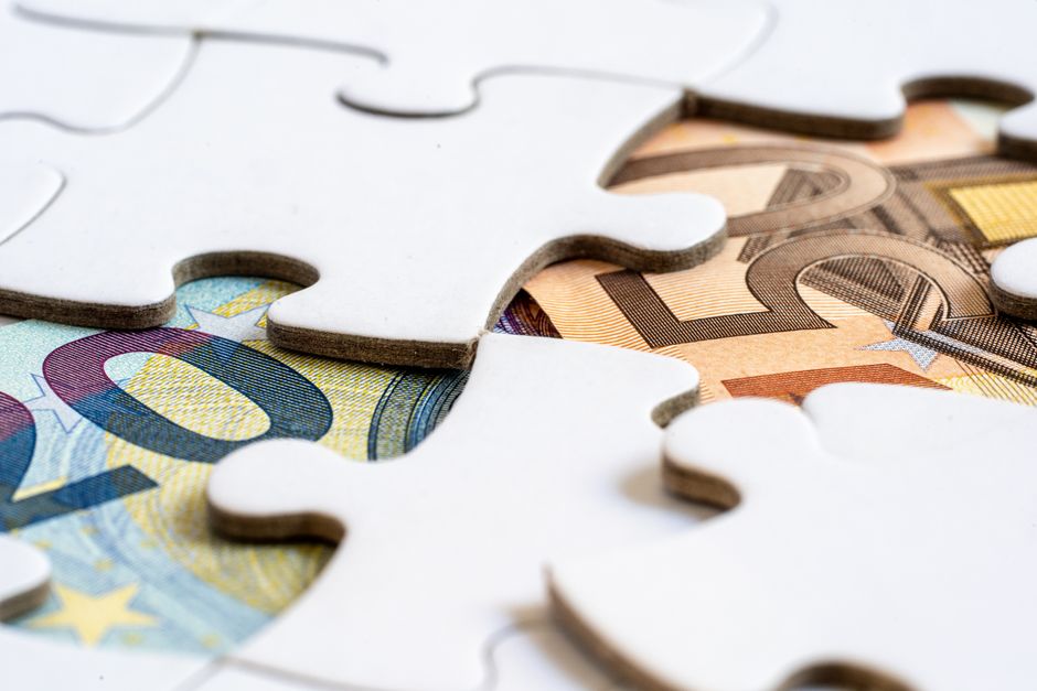 Euro banknotes partially hidden under white jigsaw puzzle pieces, symbolizing uncovered hidden costs